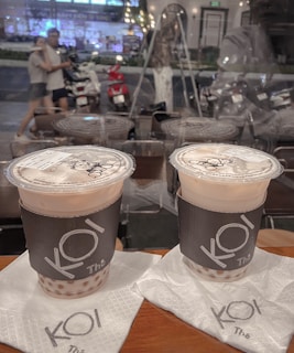 Two cups of bubble tea with branded sleeves are placed on napkins on a wooden table. In the background, a glass window reveals a blurred street scene with people walking and motorbikes parked along the roadside. The lighting appears warm and ambient.