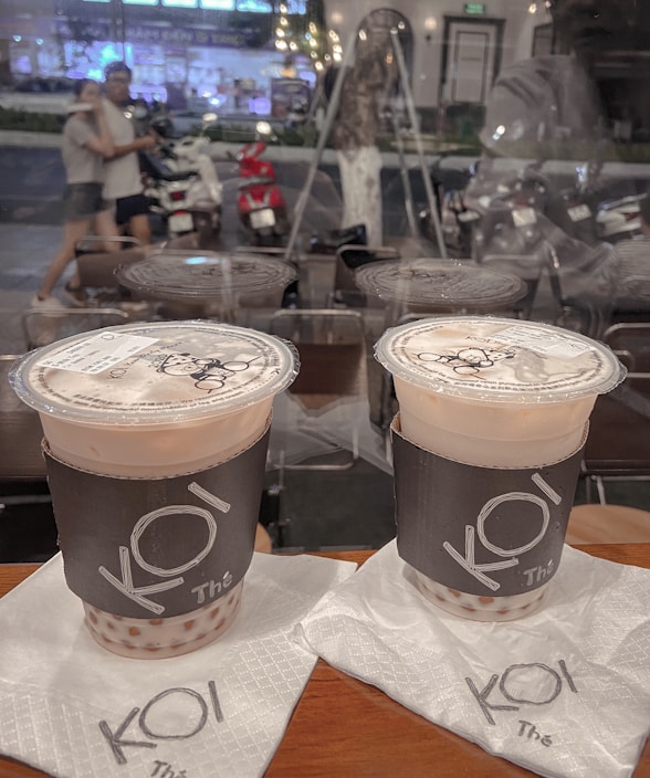 Two cups of bubble tea with branded sleeves are placed on napkins on a wooden table. In the background, a glass window reveals a blurred street scene with people walking and motorbikes parked along the roadside. The lighting appears warm and ambient.