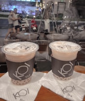 Two cups of bubble tea with branded sleeves are placed on napkins on a wooden table. In the background, a glass window reveals a blurred street scene with people walking and motorbikes parked along the roadside. The lighting appears warm and ambient.