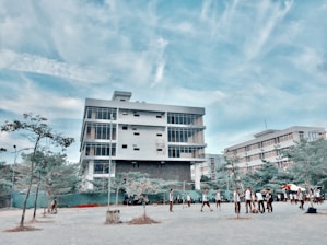 A modern, multi-story building with large windows stands prominently under a blue sky with scattered clouds. In front of the building, a group of people, possibly students, are gathered and walking around a paved area. Sparse trees and some greenery are visible in the foreground.