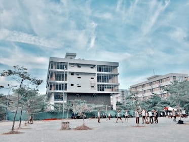 A modern, multi-story building with large windows stands prominently under a blue sky with scattered clouds. In front of the building, a group of people, possibly students, are gathered and walking around a paved area. Sparse trees and some greenery are visible in the foreground.