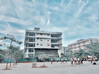 A modern, multi-story building with large windows stands prominently under a blue sky with scattered clouds. In front of the building, a group of people, possibly students, are gathered and walking around a paved area. Sparse trees and some greenery are visible in the foreground.