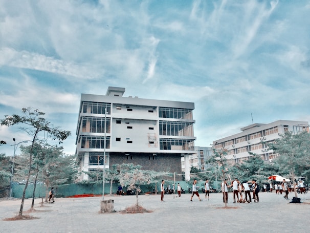 A modern, multi-story building with large windows stands prominently under a blue sky with scattered clouds. In front of the building, a group of people, possibly students, are gathered and walking around a paved area. Sparse trees and some greenery are visible in the foreground.