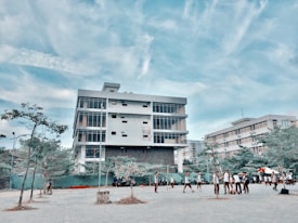 A modern, multi-story building with large windows stands prominently under a blue sky with scattered clouds. In front of the building, a group of people, possibly students, are gathered and walking around a paved area. Sparse trees and some greenery are visible in the foreground.