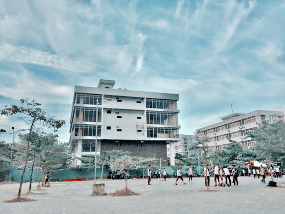 A modern, multi-story building with large windows stands prominently under a blue sky with scattered clouds. In front of the building, a group of people, possibly students, are gathered and walking around a paved area. Sparse trees and some greenery are visible in the foreground.