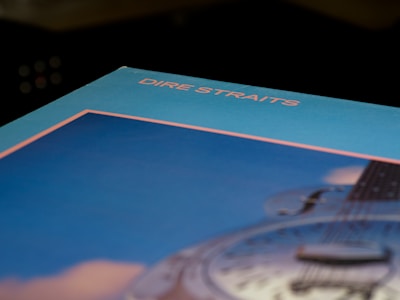 An angled view of an album cover featuring the text 'Dire Straits' in pink, set against a blue and pink background. A partial image of a guitar is visible, emphasizing musical themes.