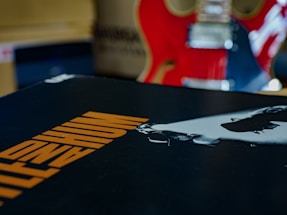 A close-up of an album cover featuring bold orange text on a black background. In the background, there is a red electric guitar with visible strings and details.