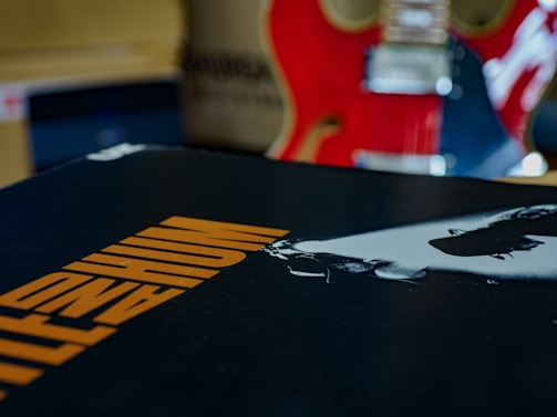 A close-up of an album cover featuring bold orange text on a black background. In the background, there is a red electric guitar with visible strings and details.