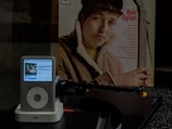 A vintage vinyl record is positioned on the turntable of a record player. To the side, a record sleeve features a young man with a guitar, wearing a brown jacket. An iPod with a white docking station is placed in the foreground, displaying a music track and album art.