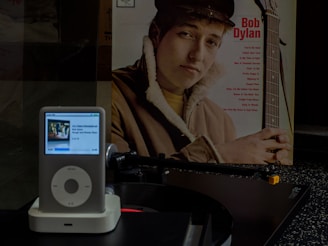 A vintage vinyl record is positioned on the turntable of a record player. To the side, a record sleeve features a young man with a guitar, wearing a brown jacket. An iPod with a white docking station is placed in the foreground, displaying a music track and album art.