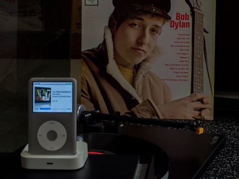 A vintage vinyl record is positioned on the turntable of a record player. To the side, a record sleeve features a young man with a guitar, wearing a brown jacket. An iPod with a white docking station is placed in the foreground, displaying a music track and album art.