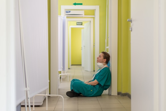 A person wearing teal medical scrubs sits on the floor with their back against a yellow wall in a corridor. The individual appears to be tired or deep in thought, with a face mask hanging around their neck. The corridor is narrow with white panels and doors, and a green arrow sign is visible on the wall.