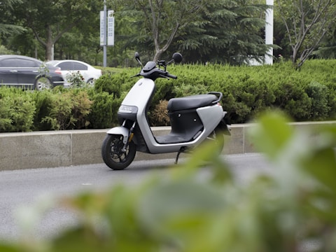 A modern electric scooter is parked on the side of a road surrounded by greenery, including bushes and trees. In the background, cars are visible driving by on the main road.