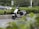 A modern electric scooter is parked on the side of a road surrounded by greenery, including bushes and trees. In the background, cars are visible driving by on the main road.