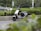 A modern electric scooter is parked on the side of a road surrounded by greenery, including bushes and trees. In the background, cars are visible driving by on the main road.