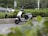 A modern electric scooter is parked on the side of a road surrounded by greenery, including bushes and trees. In the background, cars are visible driving by on the main road.