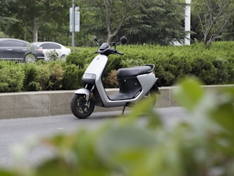 A modern electric scooter is parked on the side of a road surrounded by greenery, including bushes and trees. In the background, cars are visible driving by on the main road.