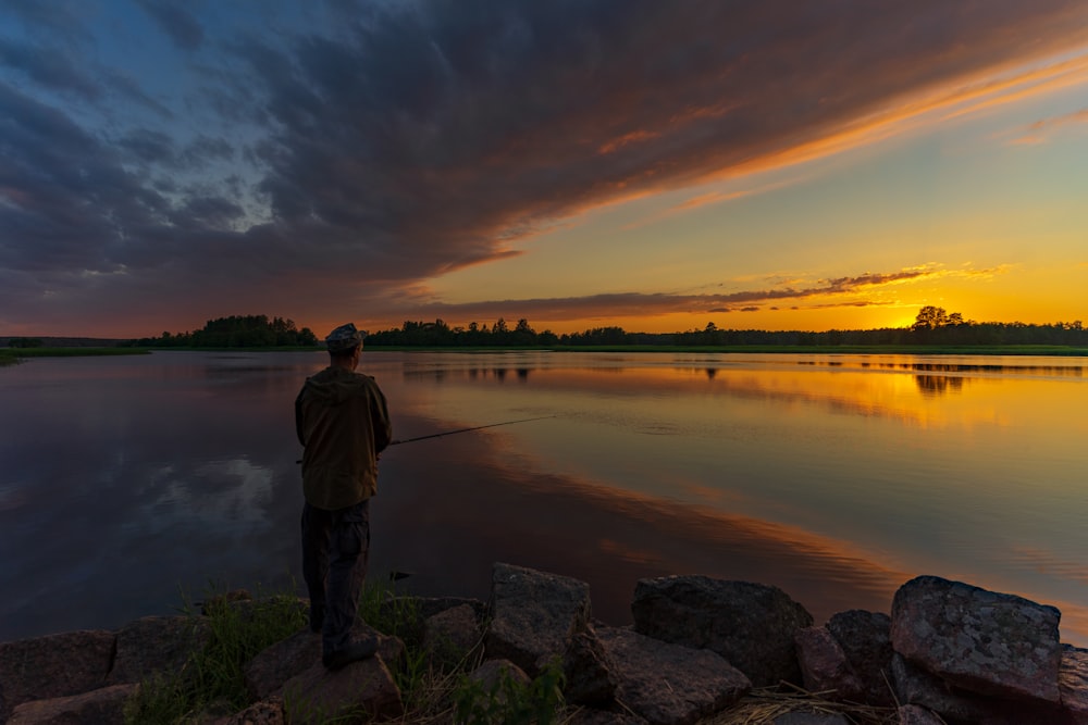 man in white jacket standing on rock near body of water during sunset