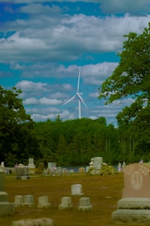 A serene cemetery with numerous gravestones scattered across a grassy area. In the background, a large wind turbine stands tall against a sky filled with fluffy clouds. Lush green trees border the cemetery, adding to the tranquil atmosphere.