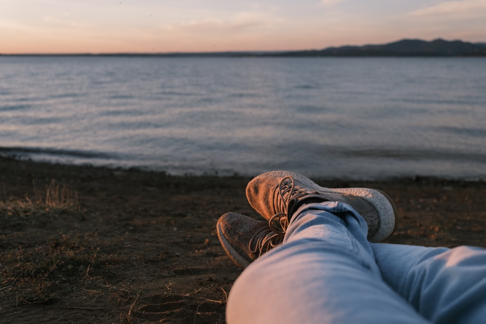 person in blue denim jeans and brown hiking shoes sitting on brown sand near body of near near near near