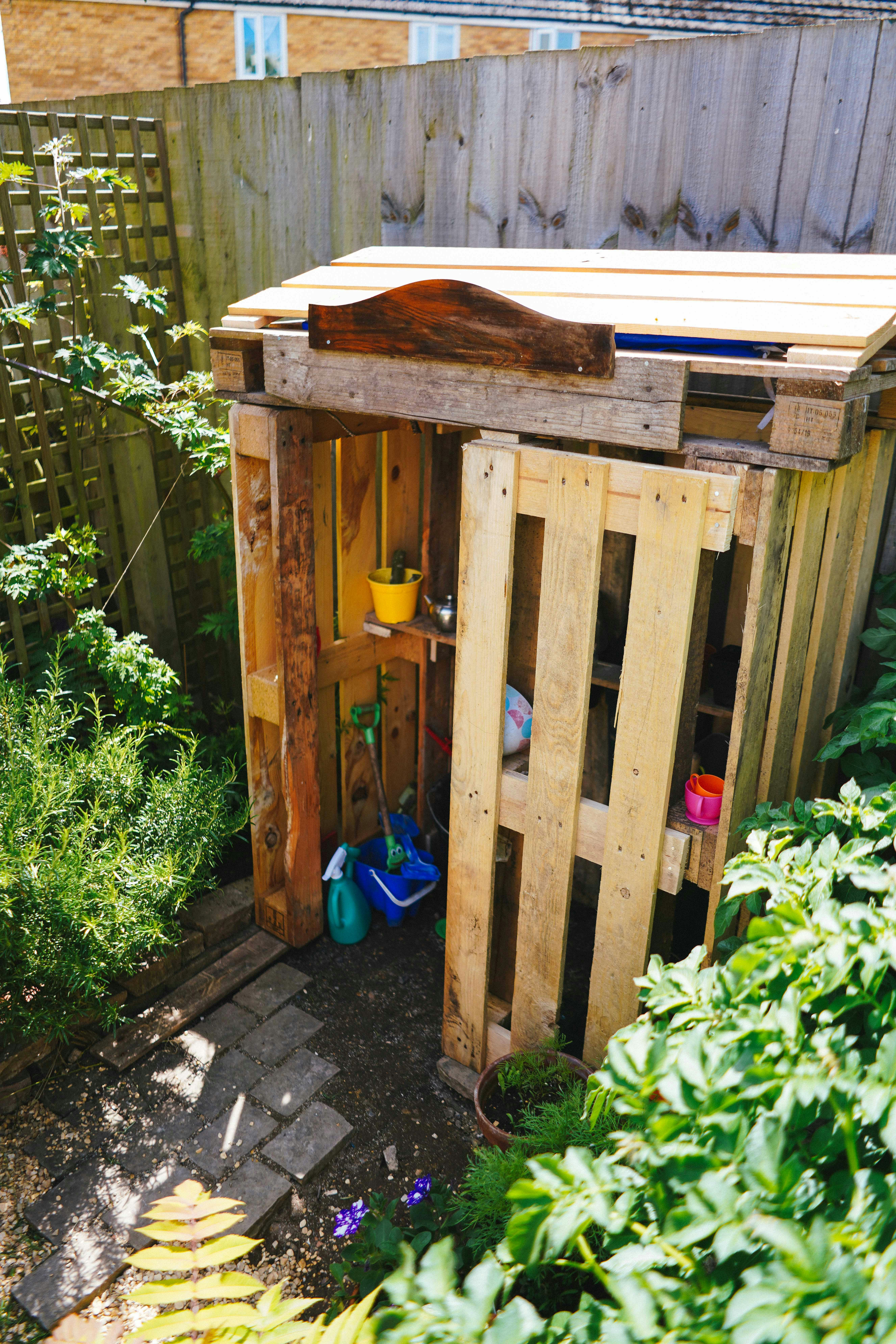 A wooden pallet shed stands in a leafy garden corner.