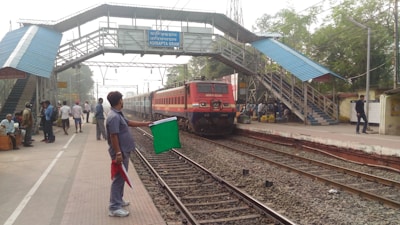 A train is approaching a station with a raised pedestrian walkway above the tracks. A railway worker is holding a green flag while standing beside the tracks, signaling the train. Several people are visible on the platform, some sitting and some walking. Buildings and trees surround the area.