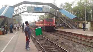 A train is approaching a station with a raised pedestrian walkway above the tracks. A railway worker is holding a green flag while standing beside the tracks, signaling the train. Several people are visible on the platform, some sitting and some walking. Buildings and trees surround the area.
