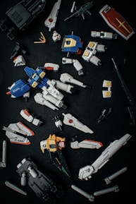 A collection of disassembled, detailed plastic model parts resembling components of a futuristic robot or mecha, laid out on a dark surface. The pieces include limbs, a torso, a shield, and various connectors, primarily in blue, white, yellow, and black.