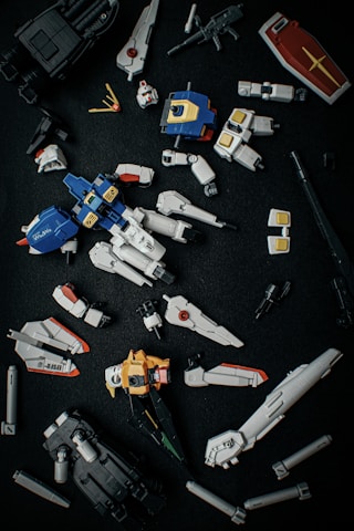 A collection of disassembled, detailed plastic model parts resembling components of a futuristic robot or mecha, laid out on a dark surface. The pieces include limbs, a torso, a shield, and various connectors, primarily in blue, white, yellow, and black.