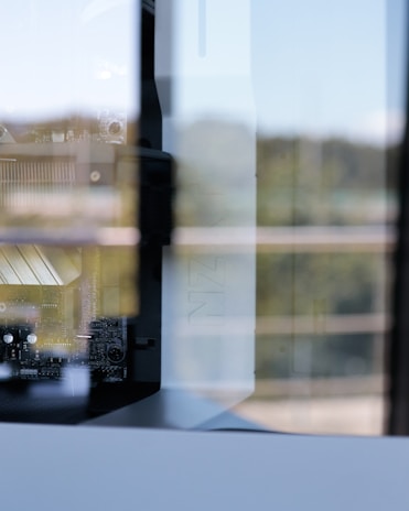 A close-up view of a computer's internal components, including a motherboard and cooling system. The image captures reflections of the surroundings on the glass panel of the computer case, adding a layer of complexity to the scene. The focus is on the intricate details of the hardware.