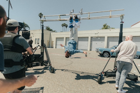 A person is hanging upside down from a rig setup, surrounded by crew members operating camera equipment. The scene appears to be a behind-the-scenes shot from a film or video production. Several people are present, including a camera operator and what seems to be assistants or support staff. The setting is an outdoor parking area near a building with multiple garage doors and an older model car is parked near the building.