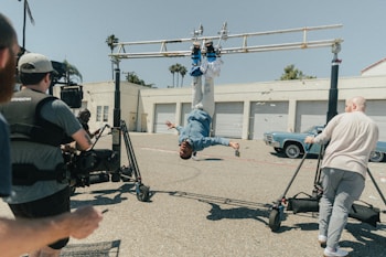 A person is hanging upside down from a rig setup, surrounded by crew members operating camera equipment. The scene appears to be a behind-the-scenes shot from a film or video production. Several people are present, including a camera operator and what seems to be assistants or support staff. The setting is an outdoor parking area near a building with multiple garage doors and an older model car is parked near the building.