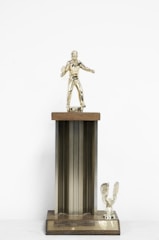 A vintage trophy featuring a golfer figurine on top of a tall, ribbed column with a wooden plaque. The figurine is positioned as if mid-swing, holding a golf club. At the base, there is a small eagle figure with its wings spread. The overall color is a mix of gold and brass tones.