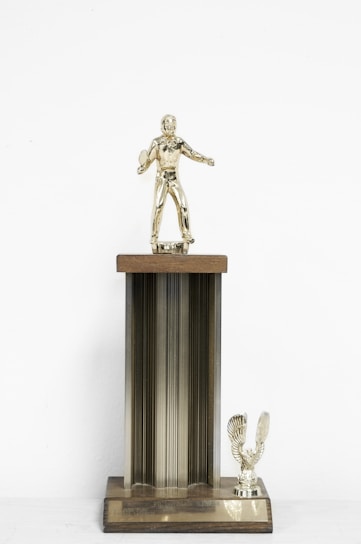 A vintage trophy featuring a golfer figurine on top of a tall, ribbed column with a wooden plaque. The figurine is positioned as if mid-swing, holding a golf club. At the base, there is a small eagle figure with its wings spread. The overall color is a mix of gold and brass tones.