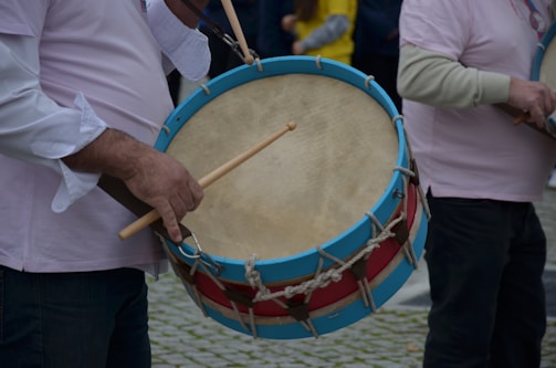 A person wearing a long-sleeved shirt holding a large traditional drum with a blue and red rim, playing it with a drumstick. Another person stands nearby, partially visible, also holding a musical instrument. The background includes blurred figures dressed in casual clothing.