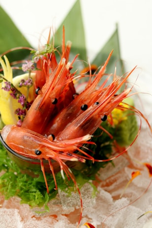 A culinary presentation featuring fresh shrimp placed in a glass container, garnished with lemon slices, green leafy vegetables, and small purple flowers. The shrimp are vibrant orange, displayed with their antennae prominently visible. The dish is arranged over a bed of crushed ice with green banana leaves in the background.
