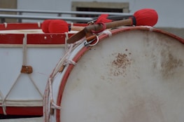 A close-up view of traditional percussion instruments featuring two large drums with red rims. The foreground highlights a drum with a partly discolored surface, while the background showcases drumsticks wrapped in red cloth resting on another drum.
