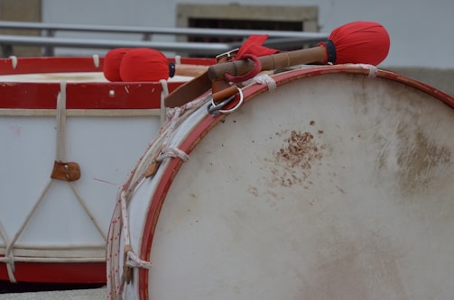 A close-up view of traditional percussion instruments featuring two large drums with red rims. The foreground highlights a drum with a partly discolored surface, while the background showcases drumsticks wrapped in red cloth resting on another drum.