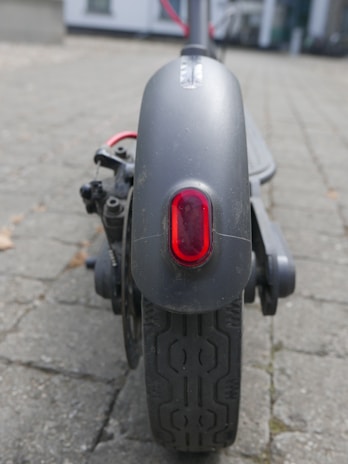 A close-up view of the rear wheel of an electric scooter, featuring a red reflector light and detailed tire tread. The background is a blurred outdoor pavement, suggesting an urban environment.