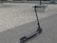 A black electric scooter is parked on a cobblestone pavement next to a gravel area. The scooter has red accents and visible handlebars. In the background, there are concrete planters and small trees.