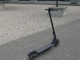A black electric scooter is parked on a cobblestone pavement next to a gravel area. The scooter has red accents and visible handlebars. In the background, there are concrete planters and small trees.
