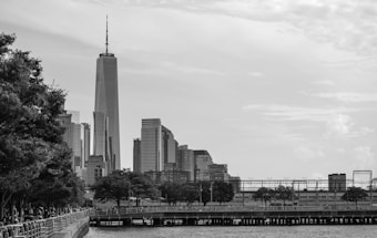 A cityscape with a prominent skyline featuring tall, modern skyscrapers. The foreground includes a waterfront with a wooden dock and trees lining a walkway. The image is rendered in black and white, highlighting architectural details and adding a classic feel.