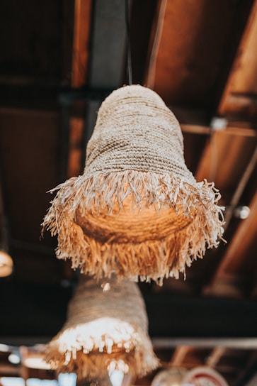 Woven lampshades made of natural fibers with a fringe, hanging from a ceiling in a cozy setting. The background is slightly blurred, suggesting a warm atmosphere with wooden elements.