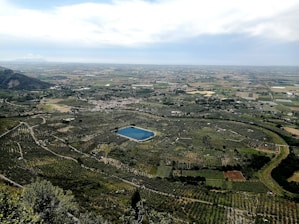 A vast landscape of agricultural fields and orchards, with a small rectangular pond located centrally in the image. Rolling hills can be seen in the background, surrounded by dense green vegetation. A few scattered buildings and patches of farmland are visible, with a distant horizon under a cloudy sky.