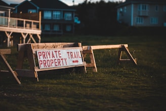 A wooden barrier with a sign that reads 'Private Property Trail' in red capital letters. The barrier stands on a grassy area with residential buildings in the background, partially illuminated by warm evening light. The atmosphere suggests a restricted or private access space.