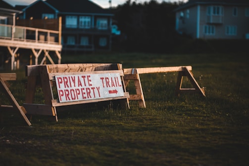 A wooden barrier with a sign that reads 'Private Property Trail' in red capital letters. The barrier stands on a grassy area with residential buildings in the background, partially illuminated by warm evening light. The atmosphere suggests a restricted or private access space.