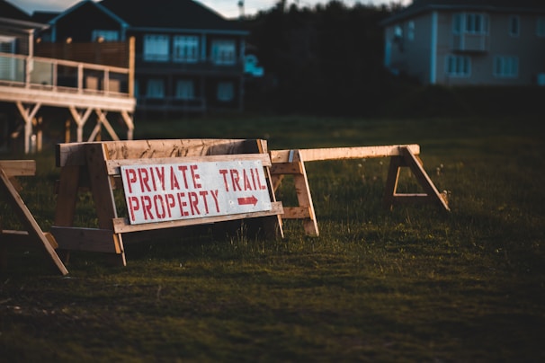 A wooden barrier with a sign that reads 'Private Property Trail' in red capital letters. The barrier stands on a grassy area with residential buildings in the background, partially illuminated by warm evening light. The atmosphere suggests a restricted or private access space.
