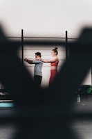 A man and a woman are exercising together, with the woman extending her arms in a workout space. There are blurred foreground elements creating a frame around them. They appear to be using resistance bands, and there is a water bottle on the floor.