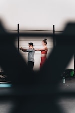 A man and a woman are exercising together, with the woman extending her arms in a workout space. There are blurred foreground elements creating a frame around them. They appear to be using resistance bands, and there is a water bottle on the floor.