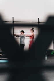 A man and a woman are exercising together, with the woman extending her arms in a workout space. There are blurred foreground elements creating a frame around them. They appear to be using resistance bands, and there is a water bottle on the floor.